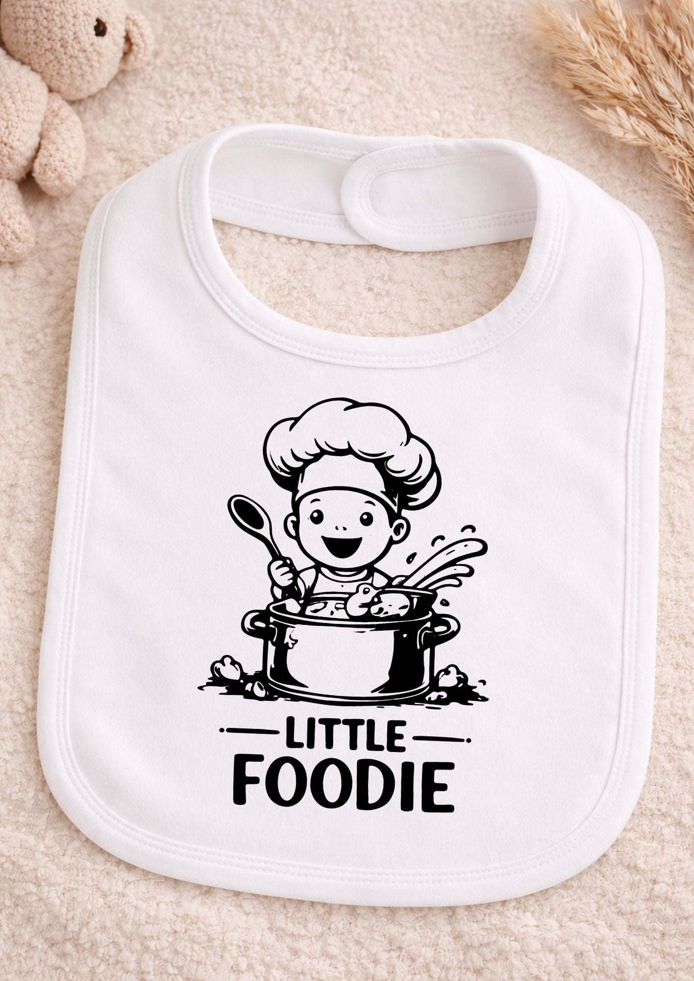 White bib with 'Little Foodie' design featuring a chef cartoon on a beige surface.