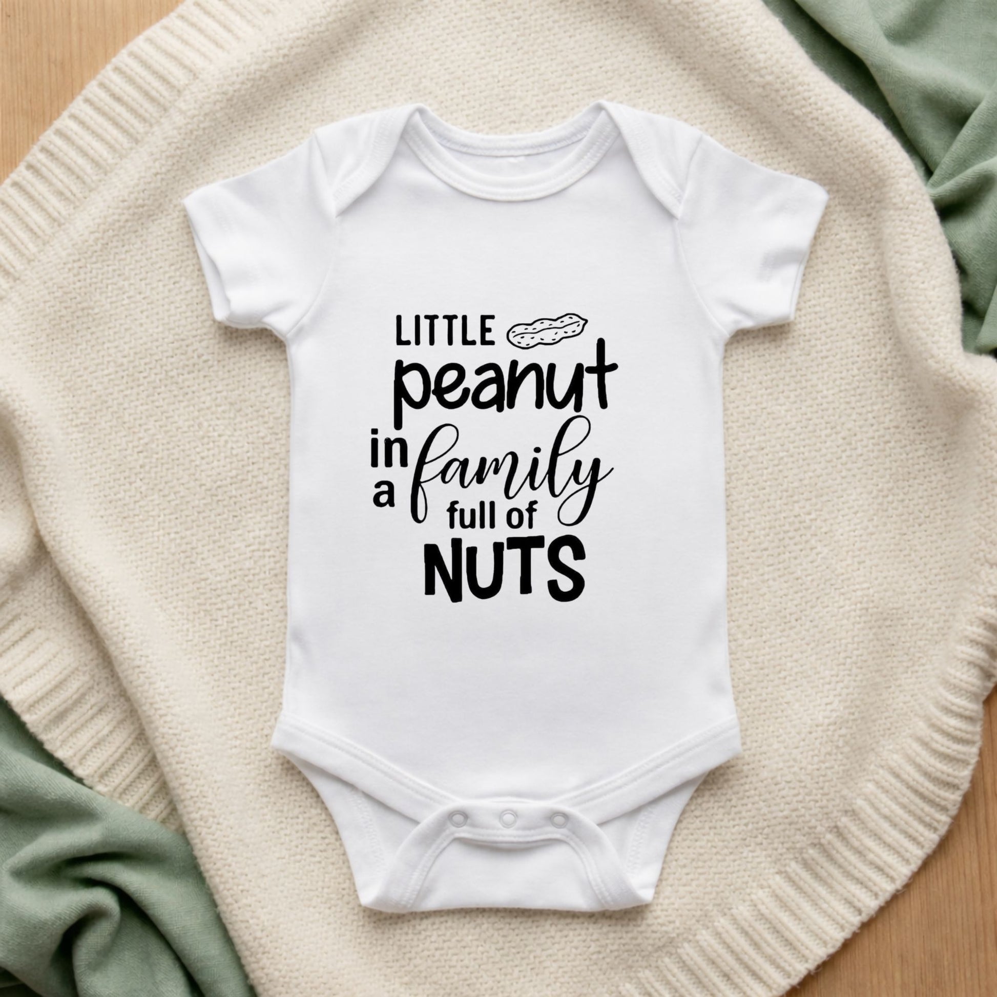 White baby onesie with humorous text design on a textured surface Little Peanut In A Family Full Of Nuts Baby Bodysuit