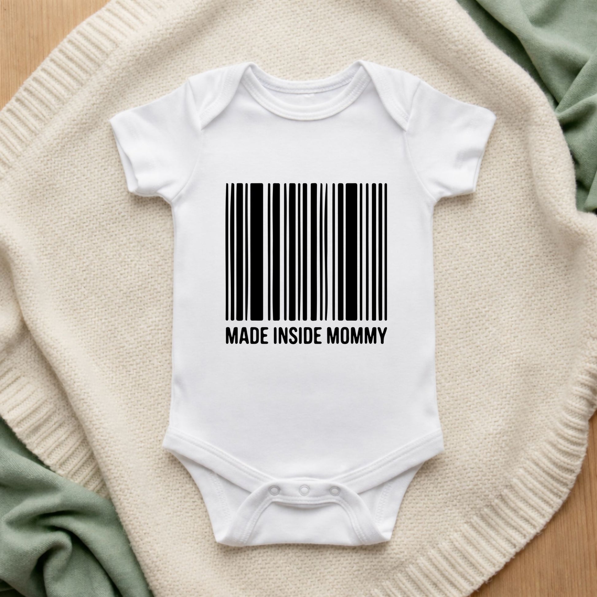 White baby onesie with a barcode and 'MADE INSIDE MOMMY' text on a textured surface.