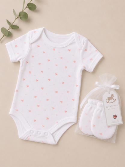 White baby bodysuit with pink heart patterns and matching white booties on a beige background.