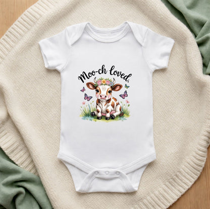 White baby onesie with a cow illustration and 'Moo-ch loved' text on a textured surface.