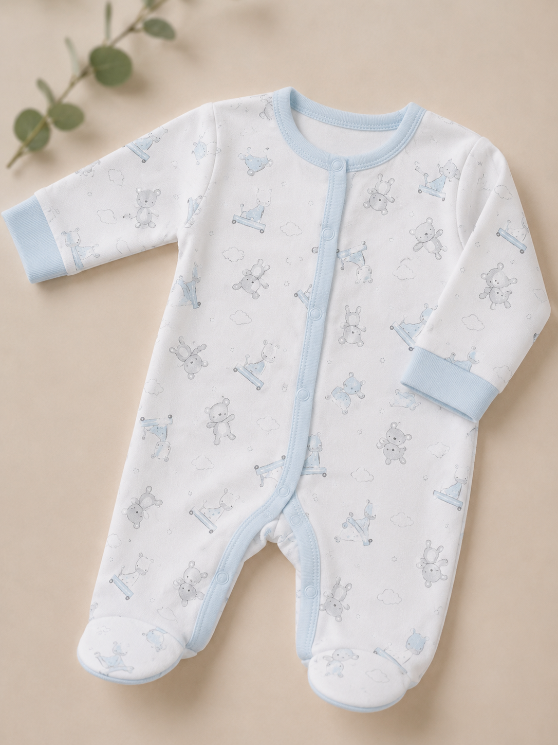 A baby long-sleeve footed sleeper in white with light blue trim and a light blue cartoon animal print, laid flat on a beige background with a small eucalyptus sprig in the corner.