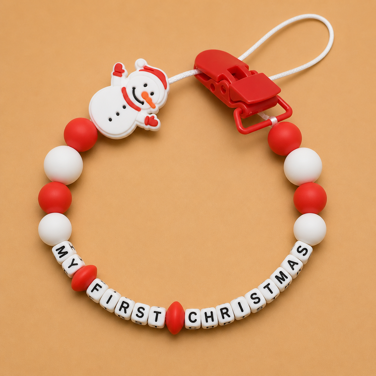 Baby dummy clip with snowman design and 'My First Christmas' text on a beige background