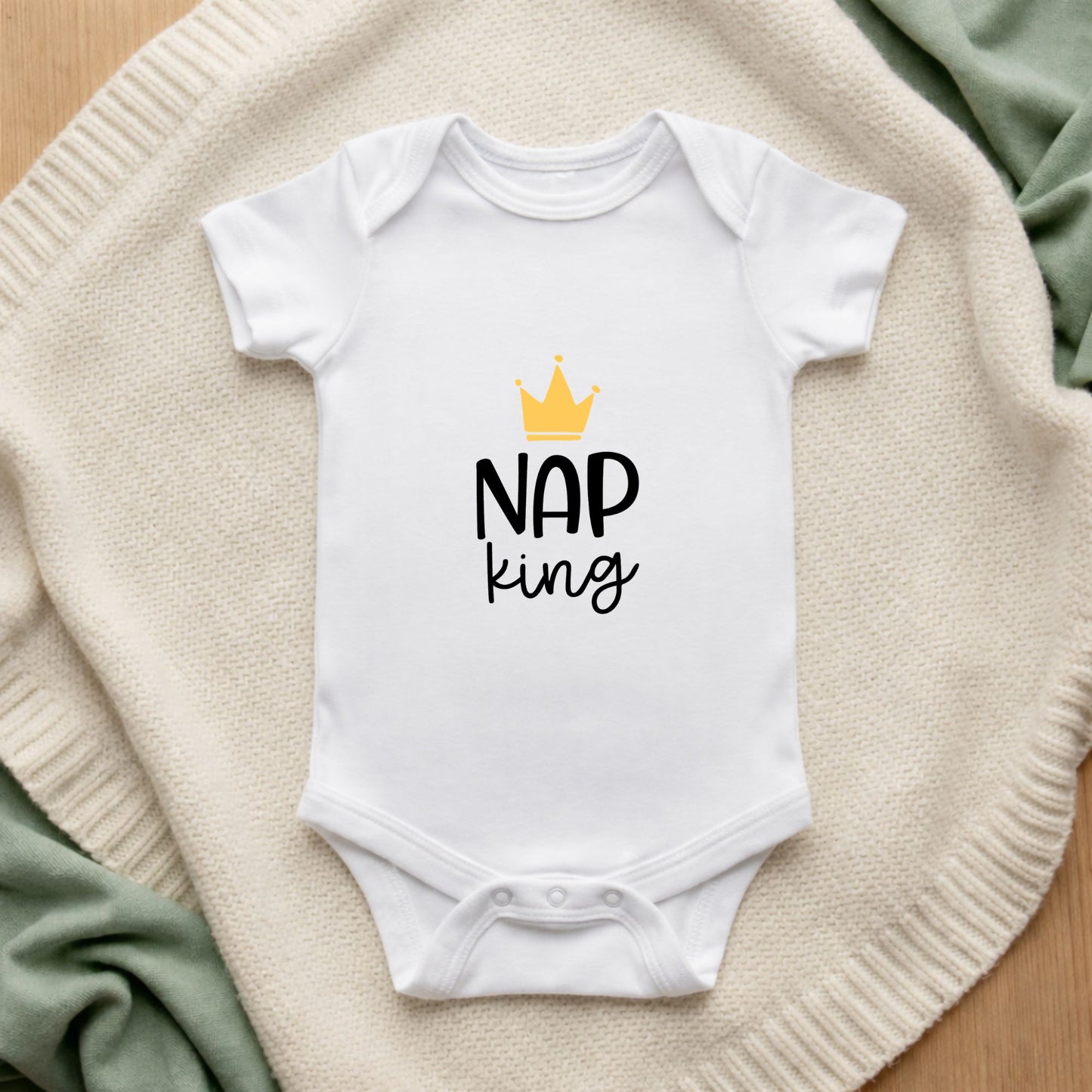 White baby onesie with 'Nap King' text and crown graphic on a textured surface.