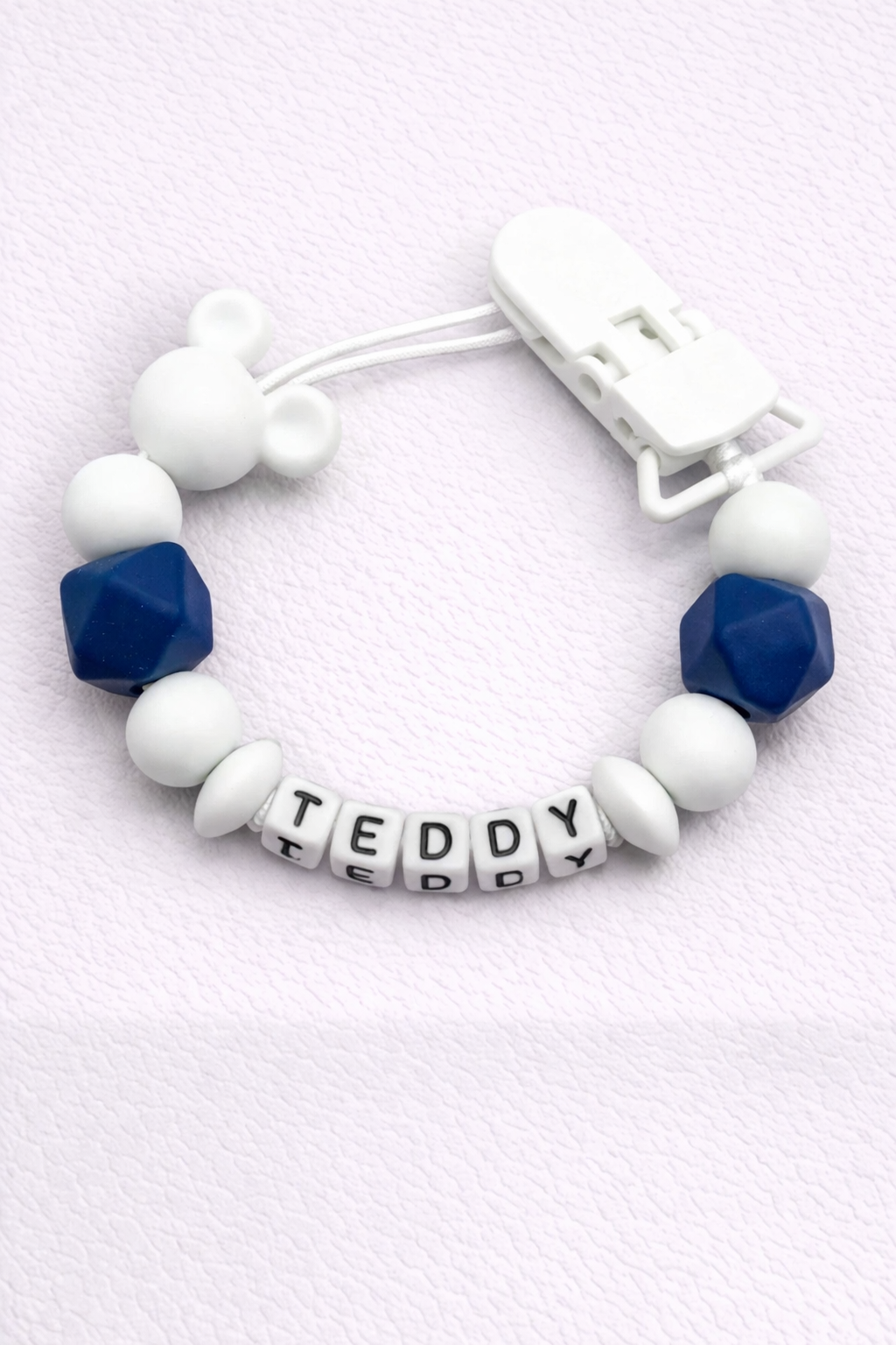 dummy clip with letter beads spelling 'TEDDY' on a light purple background