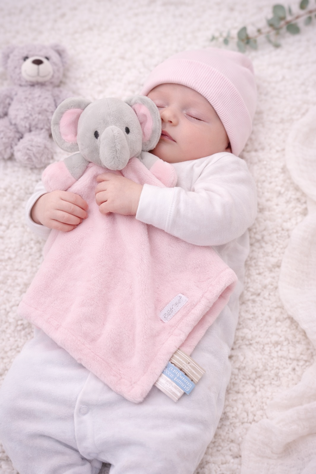 Sleeping newborn baby wearing a pink hat and white sleepsuit while cuddling a soft pink elephant comforter, photographed on a cosy cream nursery blanket with plush toys.