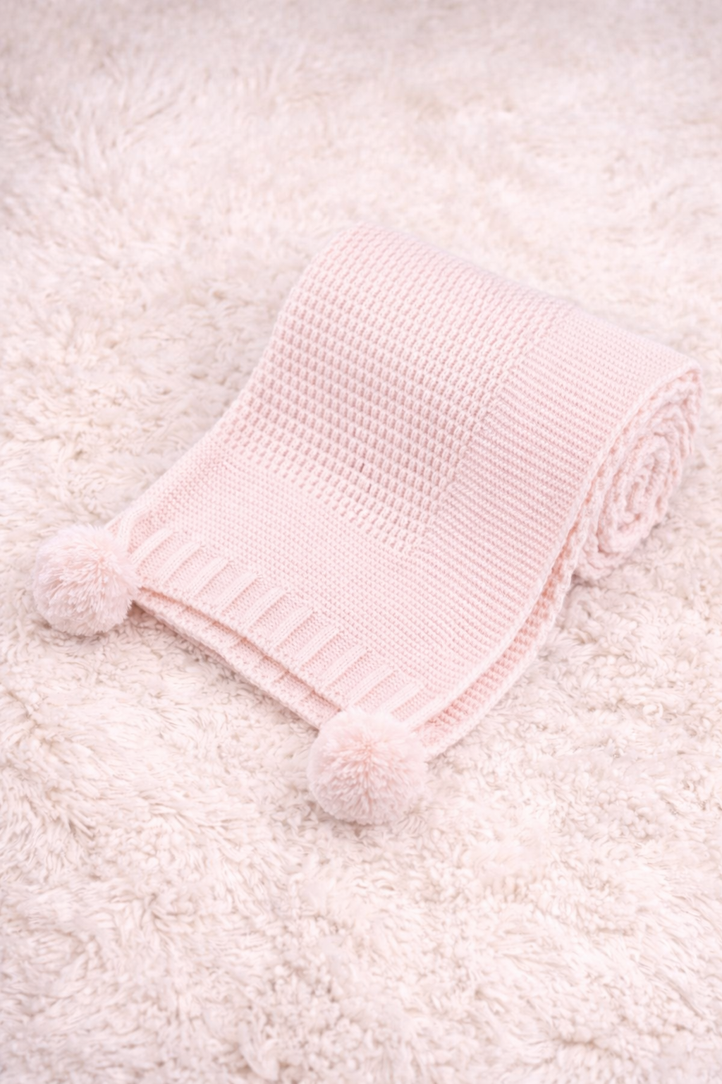 Peaceful newborn baby lying on a plush cream blanket, wrapped in a pink cable knit baby wrap with pom poms and wearing a lilac pacifier, styled for baby product photography.