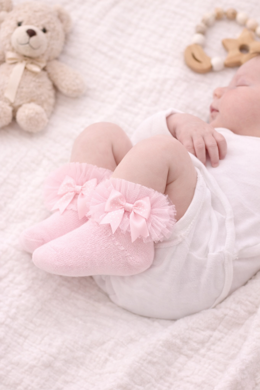 Sleeping newborn baby wearing soft pink frilly ankle socks with delicate tulle ruffles and satin bows, styled on a neutral blanket for a gentle baby photoshoot.