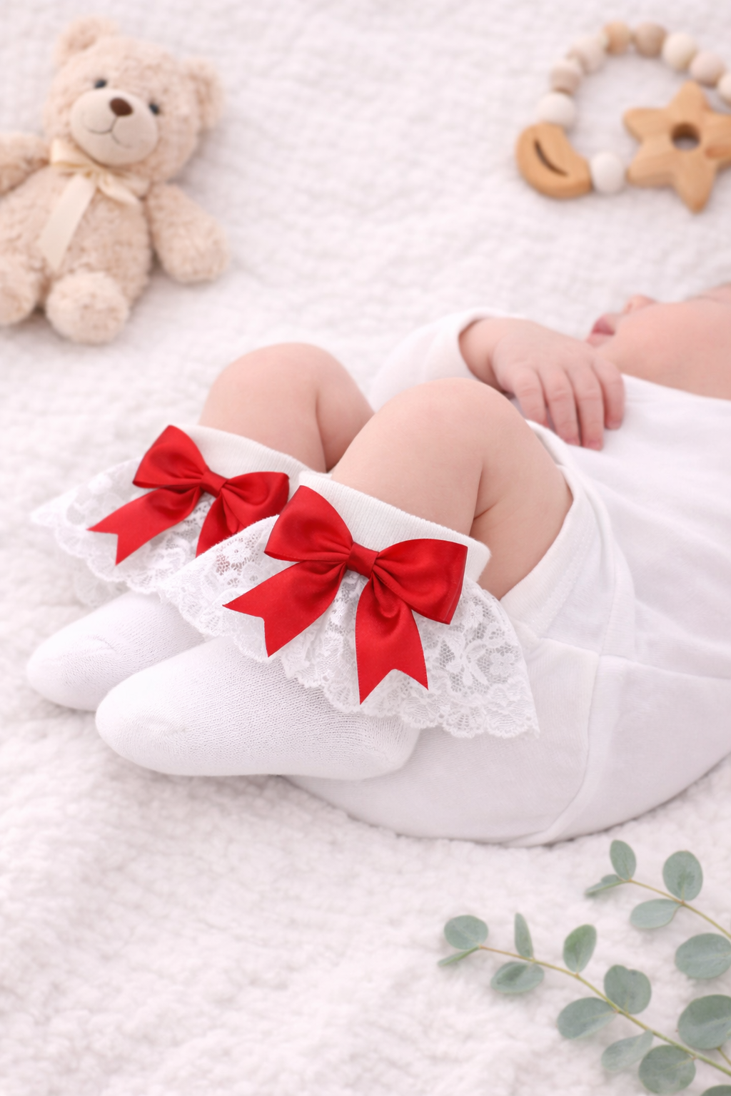 A peaceful newborn baby lies asleep on a soft neutral blanket, dressed in a white outfit and wearing elegant white lace frilly socks finished with bright red satin bows. The delicate socks are showcased on the baby’s feet, styled with a teddy bear and natural accessories for a timeless, classic baby photoshoot look.