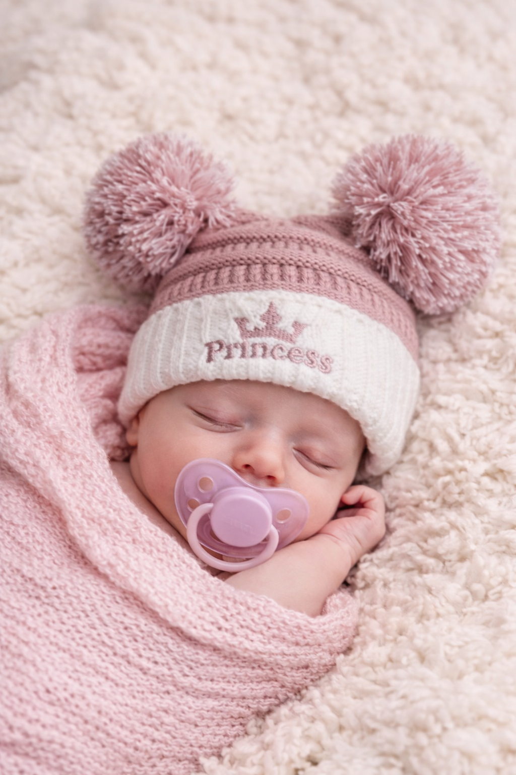 Sleeping newborn baby girl wearing a pink and cream knitted “Princess” hat with double pom poms, styled on a soft neutral blanket for a baby winter accessory mockup