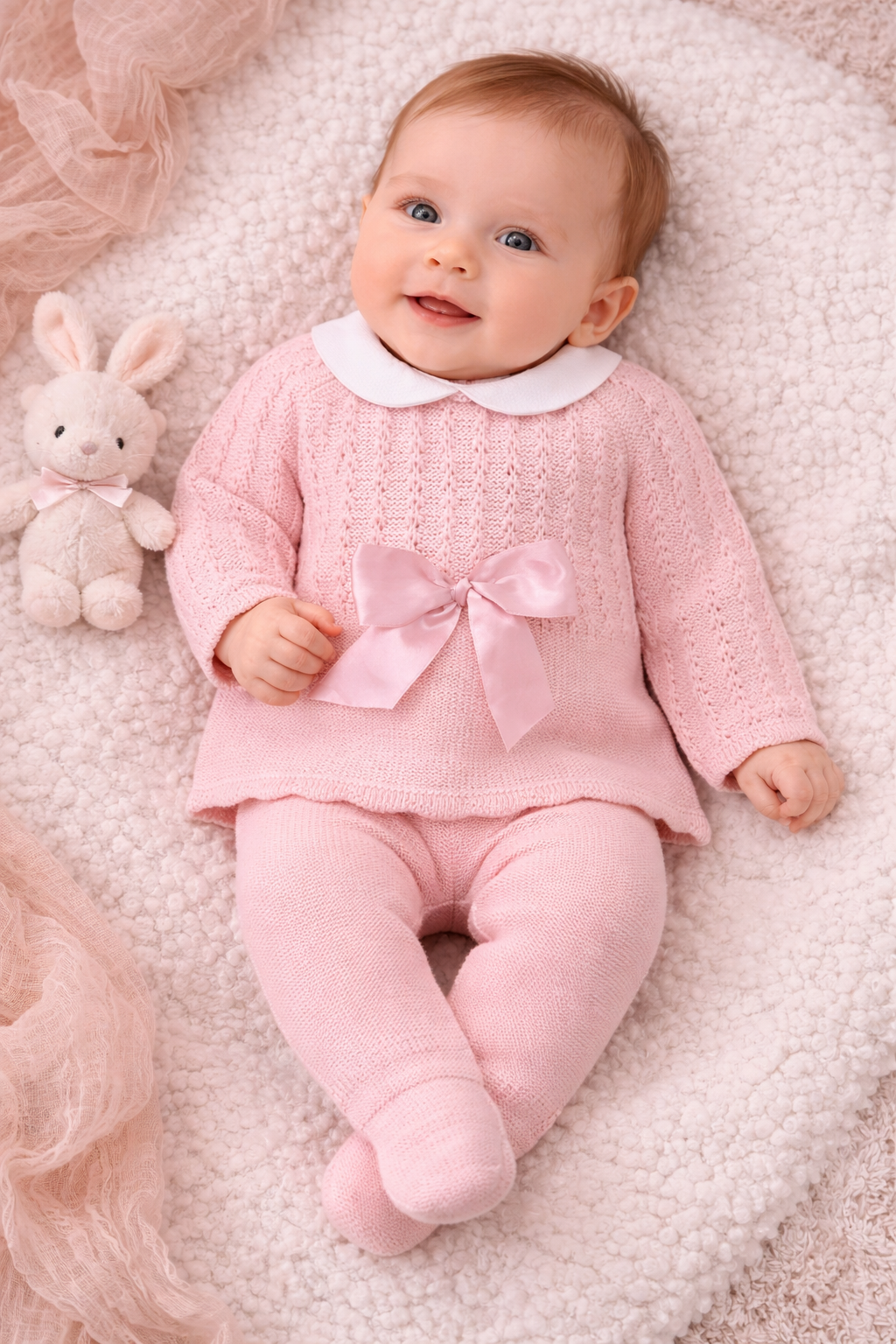 Newborn baby wearing a soft pink Spanish-style knit two-piece outfit with white Peter Pan collar and bow detail, styled in a neutral setting.