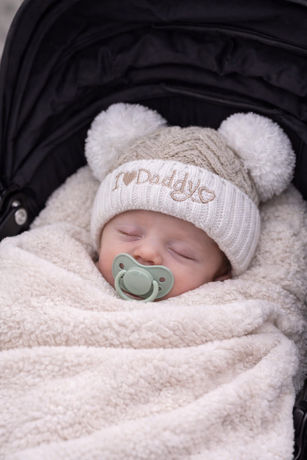 Sleeping newborn baby swaddled in a soft cream blanket inside a pram, wearing a beige chevron knit “I Love Daddy” hat with white double pom poms and a neutral dummy, styled for a cosy newborn photoshoot.