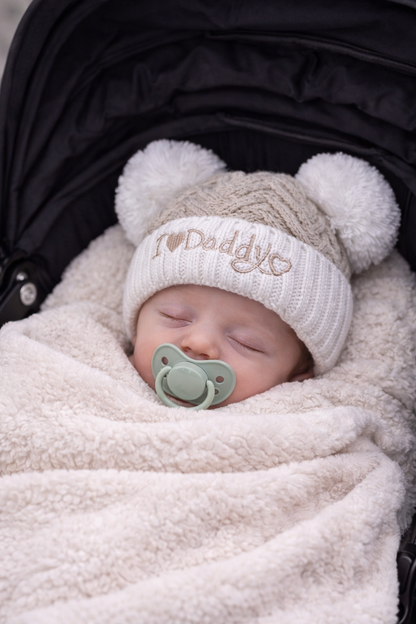 Sleeping newborn baby swaddled in a soft cream blanket inside a pram, wearing a beige chevron knit “I Love Daddy” hat with white double pom poms and a neutral dummy, styled for a cosy newborn photoshoot.