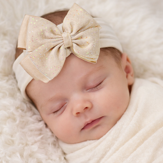 A close-up lifestyle mock-up of a peaceful newborn baby wearing a soft beige elastic headband featuring a large glitter tulle bow. The bow has subtle pastel sparkles and a classic layered design, perfectly centered on the baby’s head. The baby is sleeping on a plush neutral blanket, creating a warm, elegant, and timeless look ideal for newborn photos, baby gifts, and special occasions.