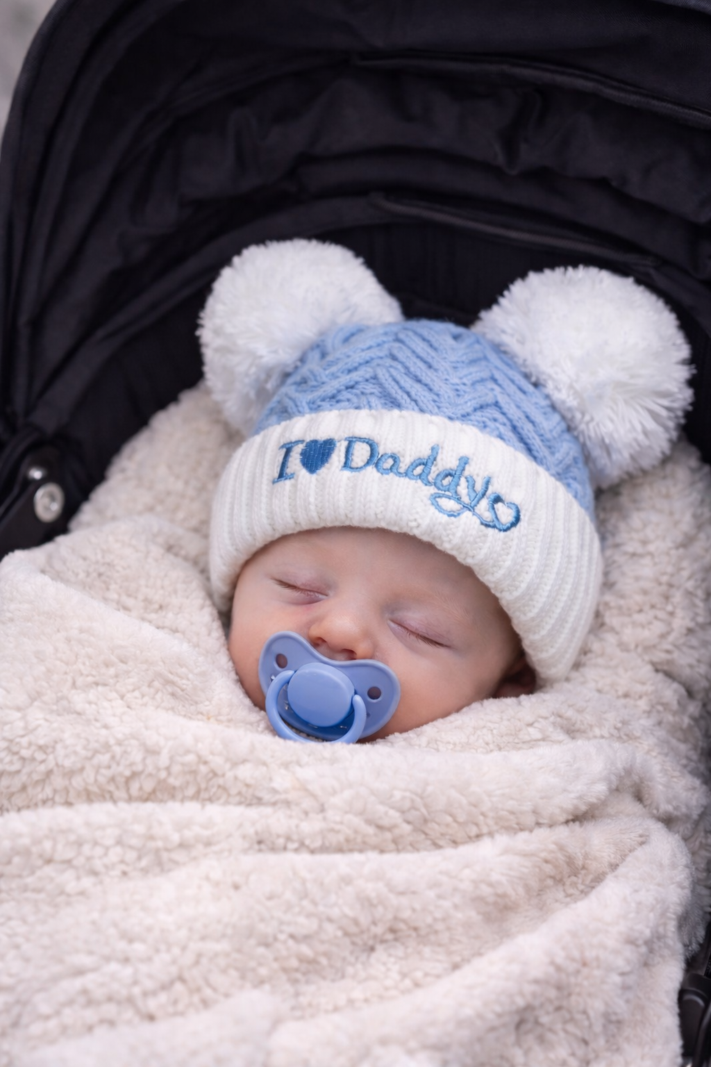 Sleeping newborn baby swaddled in a soft cream blanket, wearing a blue and white knitted “I Love Daddy” hat with double pom poms and a matching blue pacifier, resting peacefully in a pram.