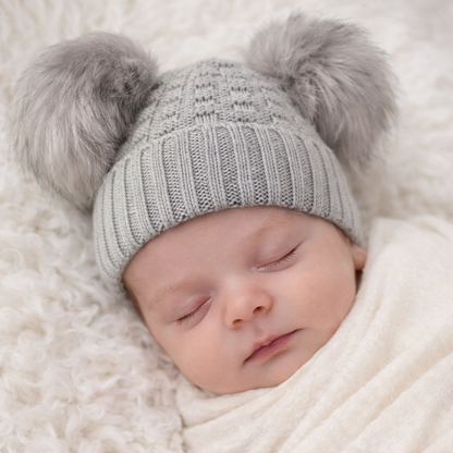 A lifestyle mock-up featuring a peacefully sleeping newborn baby wearing a soft grey knitted beanie with two fluffy pom pom details. The textured knit design fits snugly while remaining gentle on newborn skin. Styled on a plush neutral blanket with soft, natural lighting, this image highlights the hat’s cozy and timeless appeal—ideal for newborn photography, baby shower gifts, and cold-weather outings.
