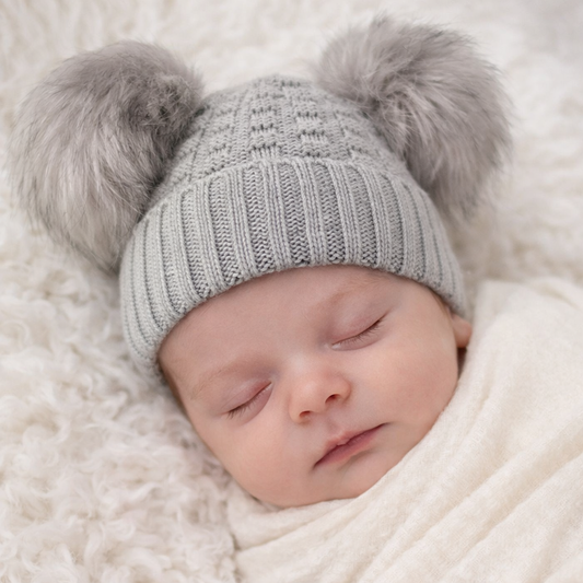 A lifestyle mock-up featuring a peacefully sleeping newborn baby wearing a soft grey knitted beanie with two fluffy pom pom details. The textured knit design fits snugly while remaining gentle on newborn skin. Styled on a plush neutral blanket with soft, natural lighting, this image highlights the hat’s cozy and timeless appeal—ideal for newborn photography, baby shower gifts, and cold-weather outings.