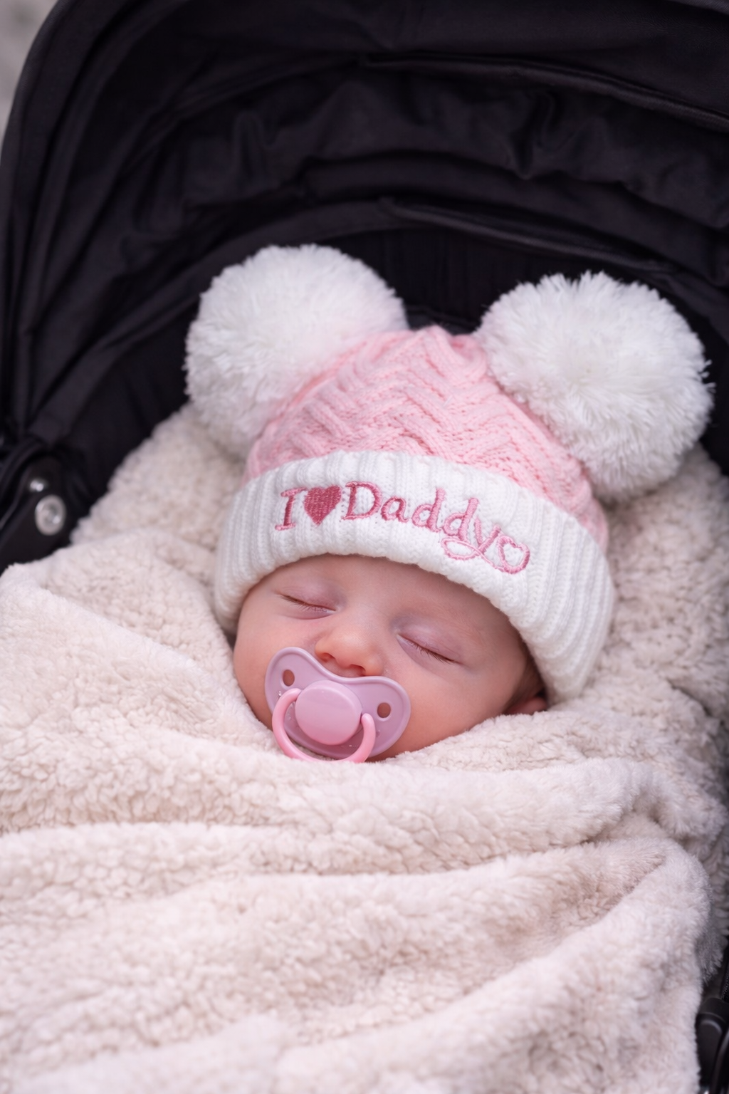 Sleeping newborn baby snug in a pram, wrapped in a soft cream blanket and wearing a pink cable knit “I Love Daddy” hat with two fluffy white pom poms and a matching pink dummy, perfect for newborn photos and baby gifts.