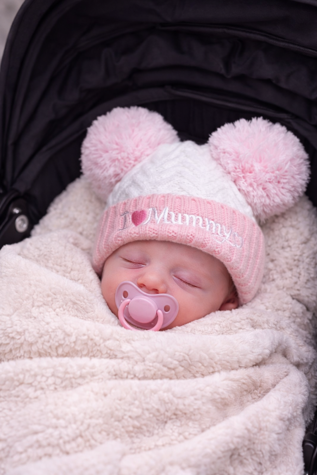 Peacefully sleeping newborn baby wrapped in a soft cream blanket inside a pram, wearing a pink and white “I Love Mummy” knitted hat with fluffy double pom poms and a matching pink dummy.