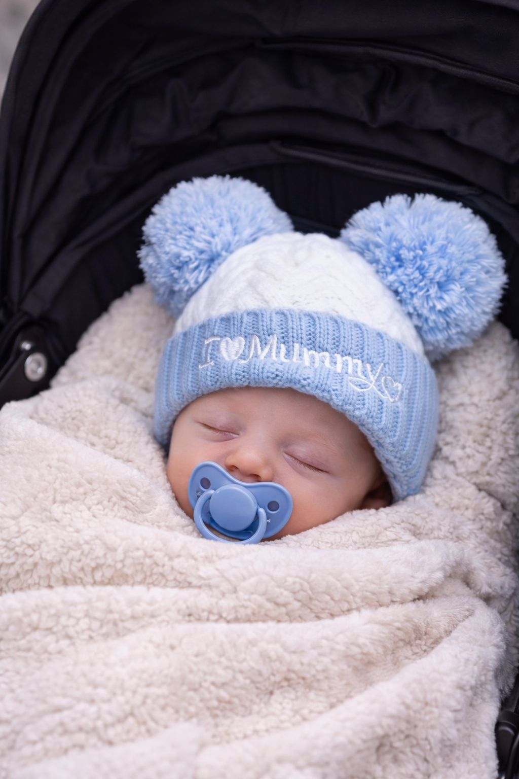 Sleeping newborn baby wrapped in a soft cream blanket inside a pram, wearing a blue and white knitted hat with double pom poms and “I Love Mummy” embroidery, styled with a matching blue dummy for a cosy winter baby mockup.