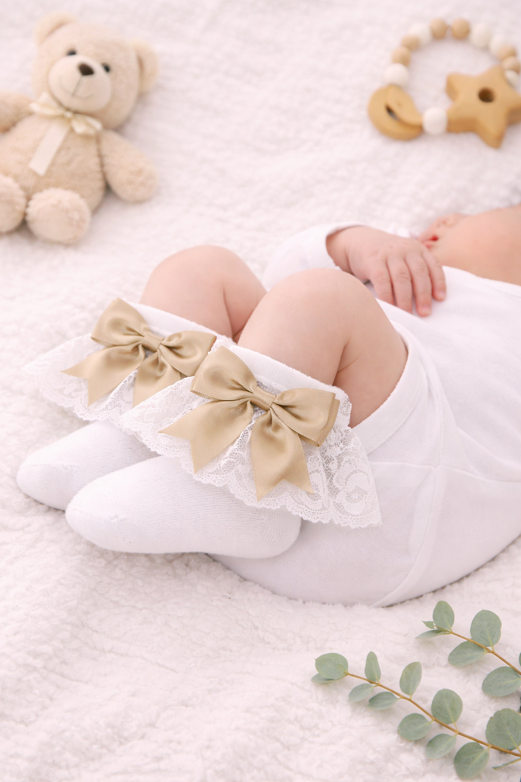 Peaceful newborn baby dressed in a white onesie, wearing elegant white lace ankle socks finished with soft champagne satin bows, styled on a cozy neutral blanket with classic nursery accessories.