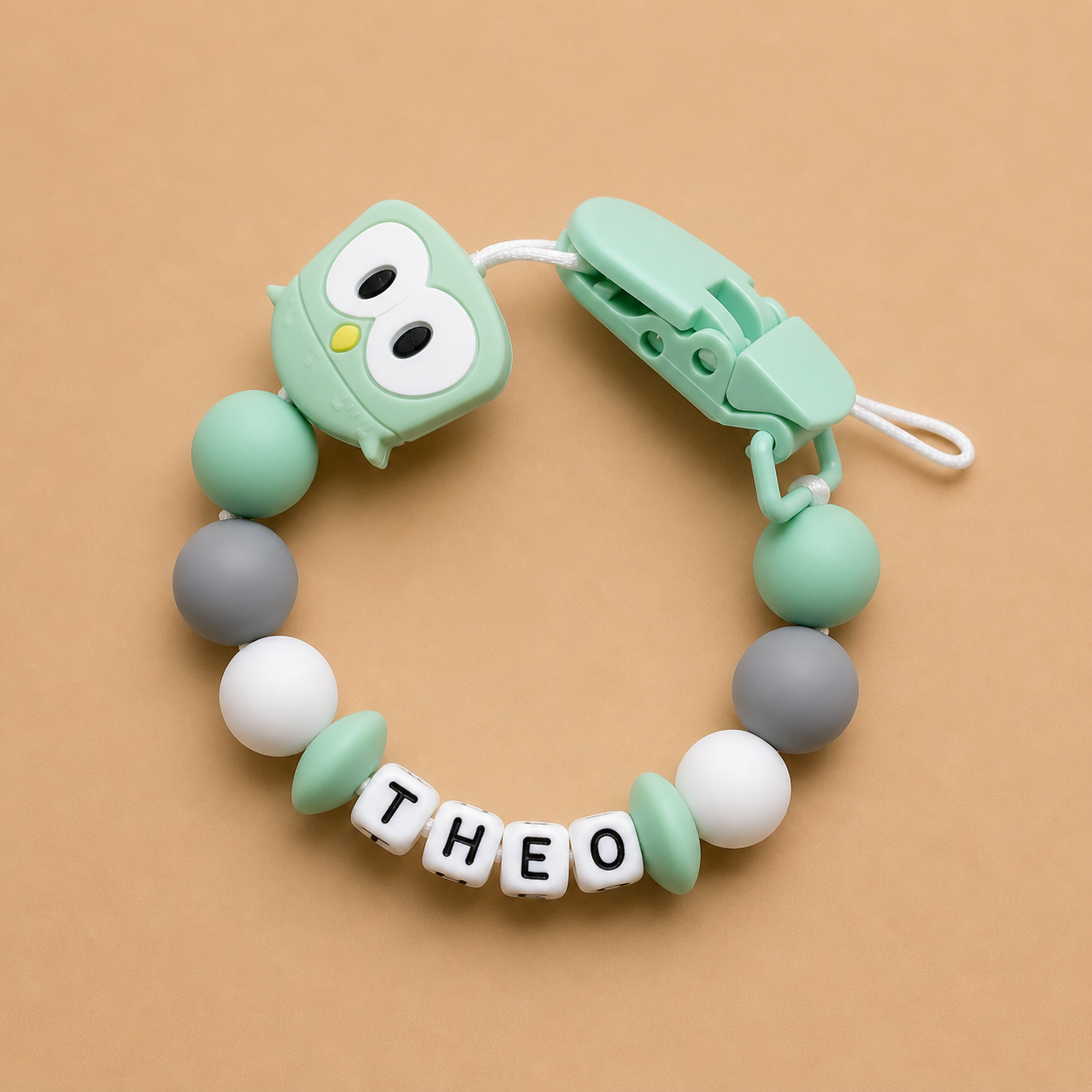 dummy clip with owl design and 'THEO' letter beads on a beige background