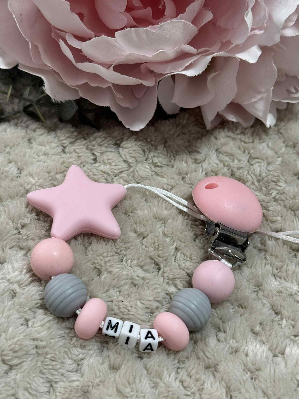 A beaded pacifier clip with pink and gray beads, a pink star, and letter beads spelling "Mia" lying on a textured Beige surface. Personalised option available