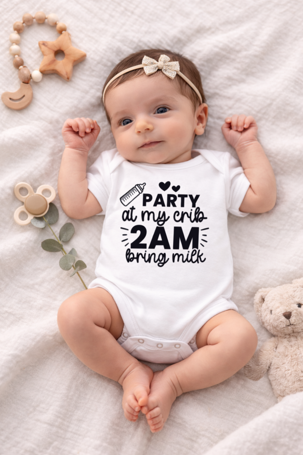 Baby wearing a onesie with text, surrounded by toys on a soft surface