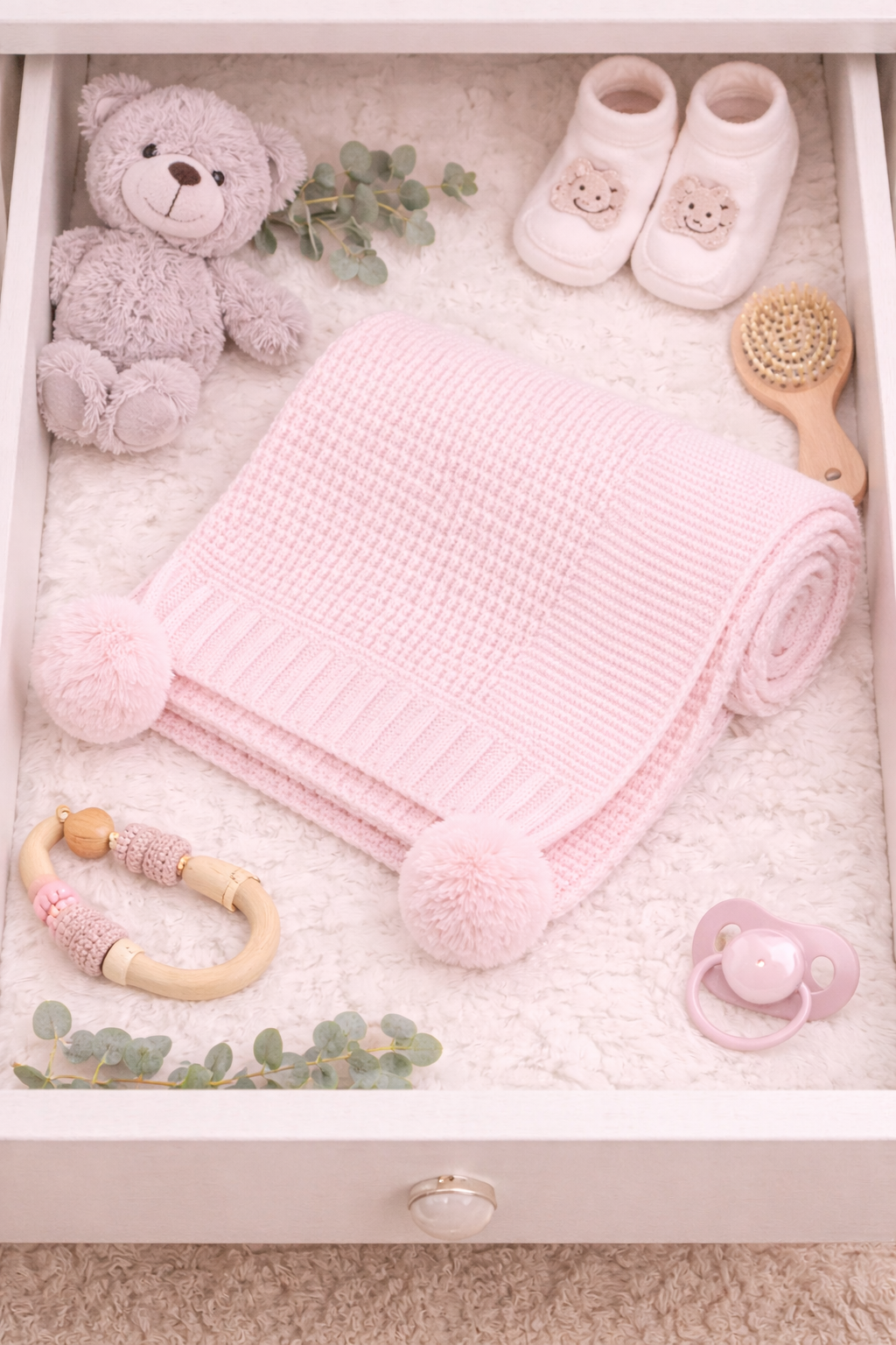 Baby care items including a pink blanket with pom-poms, teddy bear, shoes, and pacifier in a drawer.