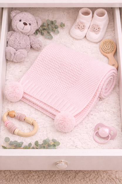 Baby care items including a pink blanket with pom-poms, teddy bear, shoes, and pacifier in a drawer.