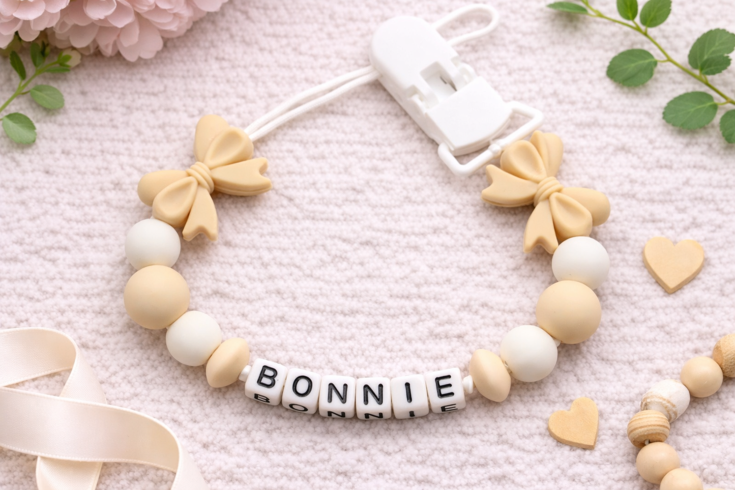Flat lay photograph of a personalised baby pacifier clip arranged in a soft, styled mock-up setting. The clip features beige bow-shaped silicone beads, cream and off-white round beads, and white letter cubes spelling “BONNIE.” It is displayed on a textured cream background with subtle decorative elements, creating a clean, neutral, and elegant product presentation.