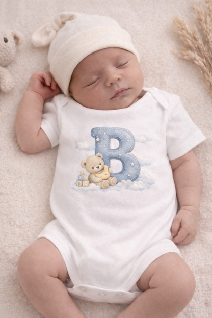 Newborn baby wearing a white onesie with a blue letter 'B' and teddy bear design, lying on a soft surface.