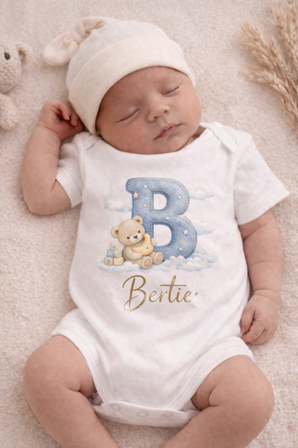 Newborn baby wearing a white onesie with a teddy bear and letter 'B' design, lying on a soft surface.