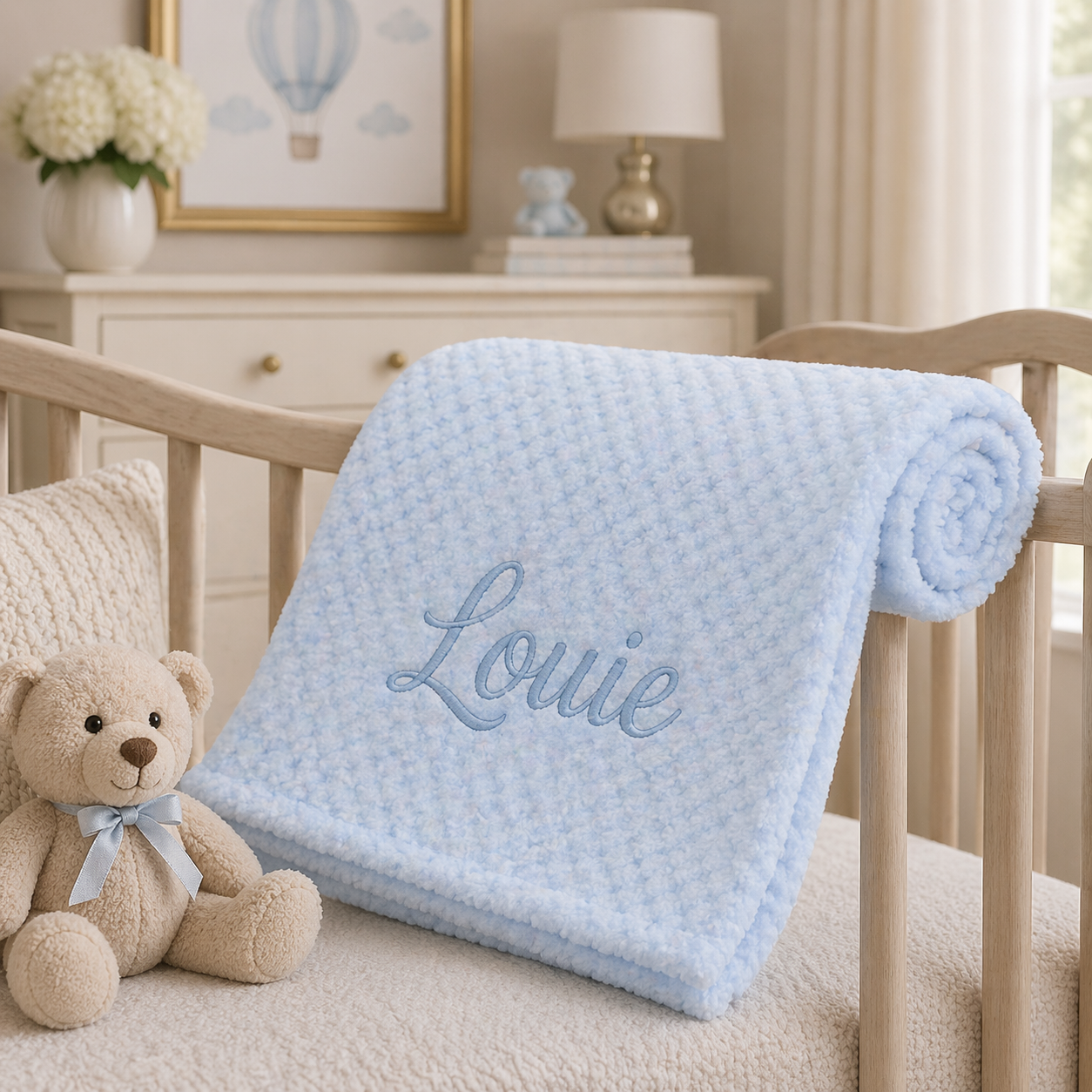 Baby crib with a blue blanket embroidered with 'Louie', a teddy bear, and decorative items in the background.