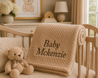 A beige textured blanket with the personalized name 'Baby McKenzie' embroidered in dark brown script, draped over a wooden crib next to a plush teddy bear in a softly decorated nursery.