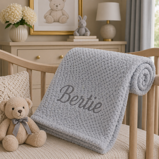 Gray blanket with 'Bertie' embroidered on a wooden crib in a nursery setting.