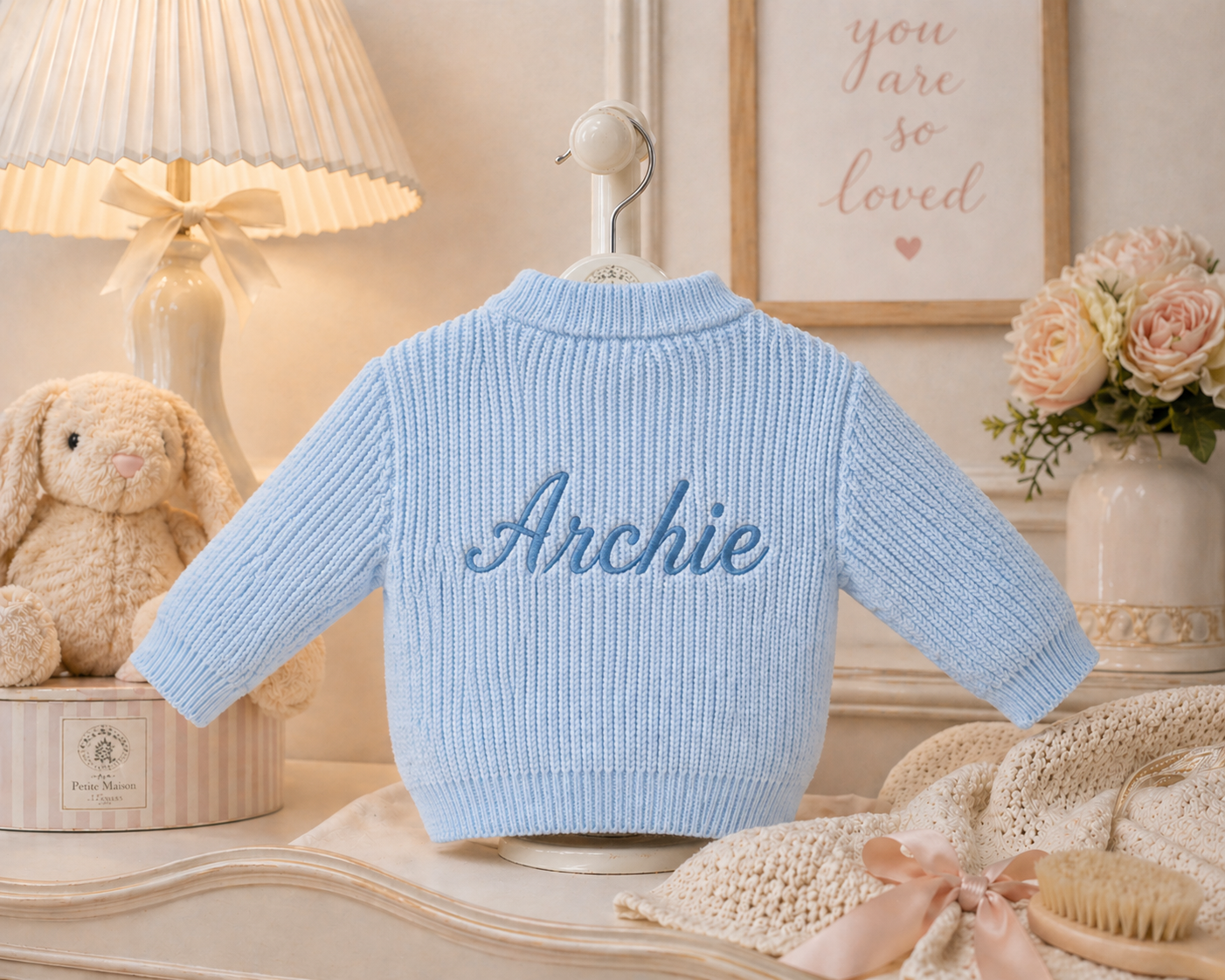 Light blue baby sweater with 'Archie' embroidered on it, placed on a decorative surface with a lamp and flowers in the background.