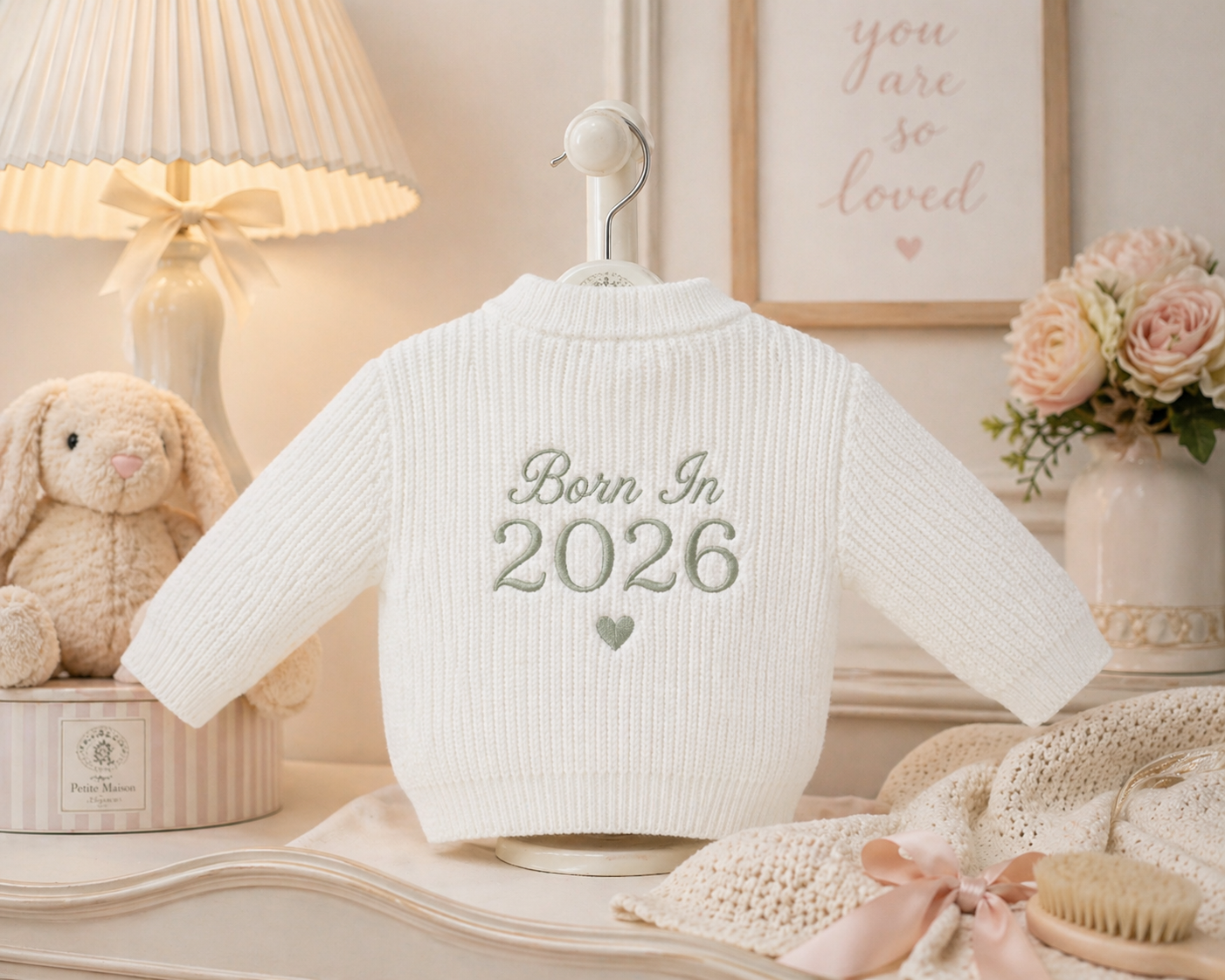 White baby sweater with 'Born In 2026' text on a hanger in a nursery setting.