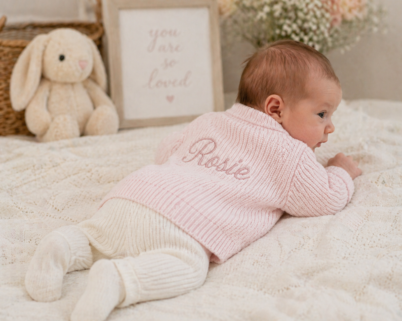 Baby in a pink cardigan with 'Rosie' embroidered, lying on a soft surface with a plush toy and framed picture in the background.
