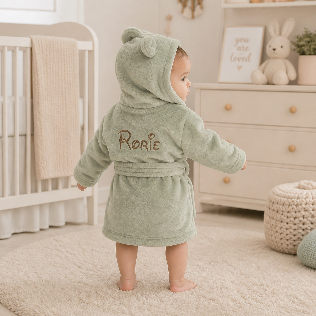 Baby in a green hooded robe with 'Rorie' embroidered in a nursery setting.