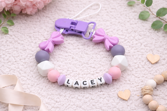 Personalised baby pacifier clip with purple, pink and grey silicone beads spelling the name Lacey, styled on a soft cream textured flat lay background with flowers and wooden heart details.