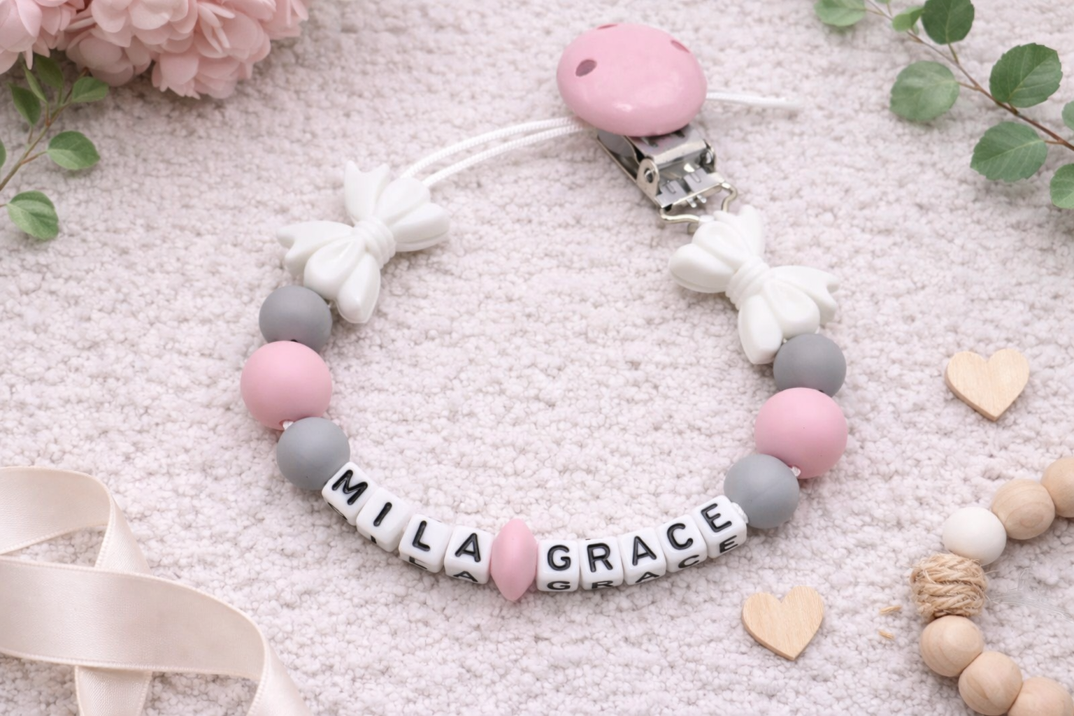 Personalised baby pacifier clip in pink, grey and white silicone beads spelling “Mila Grace” displayed on a soft neutral flat lay background with flowers and wooden hearts.