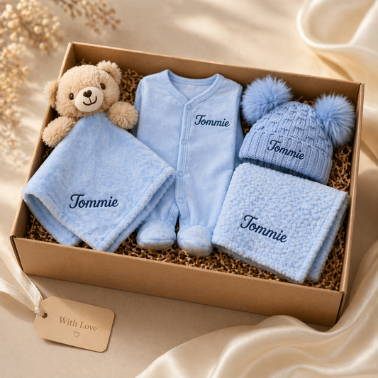 Baby gift set with blue outfit, hat, blanket, and teddy bear in a box on a beige background.