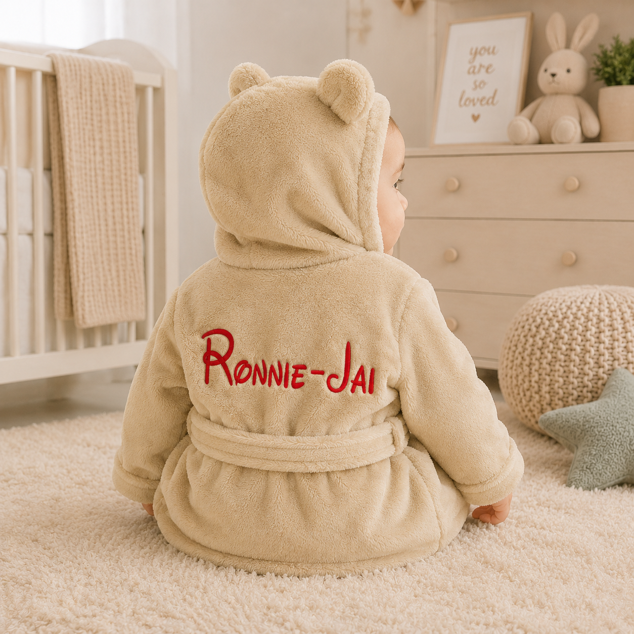 Child wearing a beige robe with 'RONNIE-JAI' embroidered on the back, standing in a nursery.