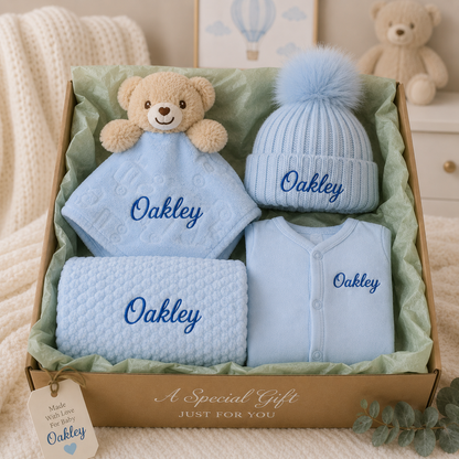 Personalised baby gift box in blue with embroidered babygrow, pom pom hat, bubble blanket and teddy bear comforter, presented in a brown gift box with tissue paper.