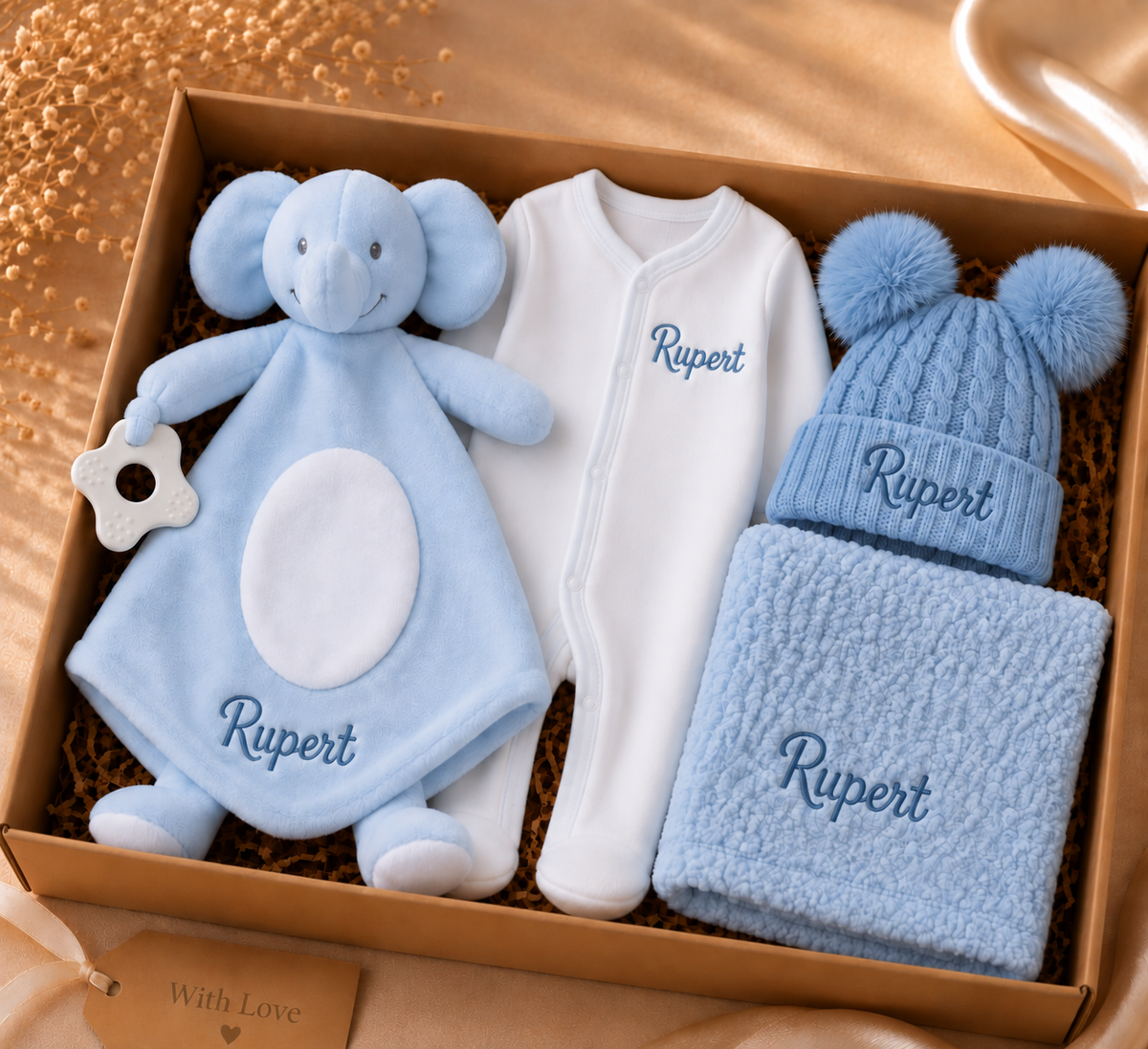 A personalized baby gift set in a brown box, featuring a light blue elephant security blanket, a white footless sleeper with 'Rupert' embroidered on the chest, a light blue knit beanie hat with pom-poms, and a matching light blue textured scarf, all with 'Rupert' embroidered in blue. The box is filled with brown paper shred and sits on a warm beige surface.