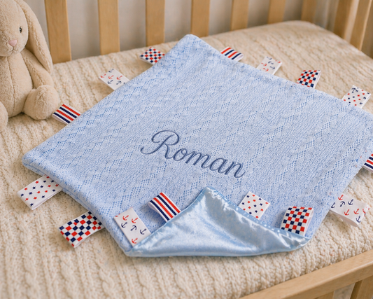 Blue baby blanket with embroidered name 'Roman' on a crib with colorful flags.