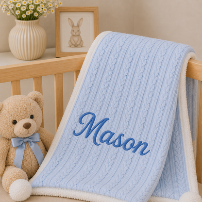A light blue cable-knit baby blanket with a white sherpa trim and the name 'Mason' embroidered in blue script, draped over a wooden crib next to a teddy bear and a framed bunny picture.