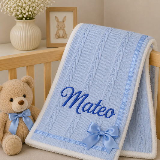 A light blue and white fleece baby blanket with a cable-knit pattern, personalized with the name 'Mateo' in blue embroidery, a light blue satin bow, and a matching ribbon trim. The blanket is draped over a wooden crib next to a plush teddy bear wearing a matching blue ribbon bow.