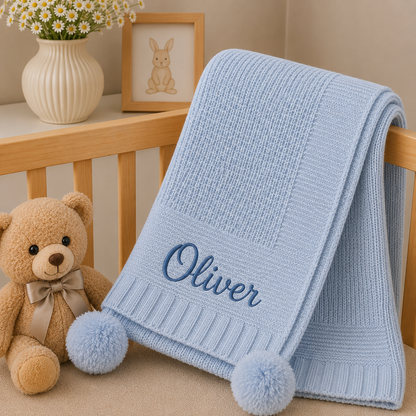 A light blue knitted baby blanket with two fluffy pom-poms and the name 'Oliver' embroidered in navy script, draped over a wooden crib next to a teddy bear and a vase of daisies.